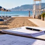 Calcolatrice, penna e documenti di progetto su un tavolo in primo piano con uno stabilimento balneare e ombrelloni sullo sfondo per il calcolo degli indennizzi dei beni ammortizzati