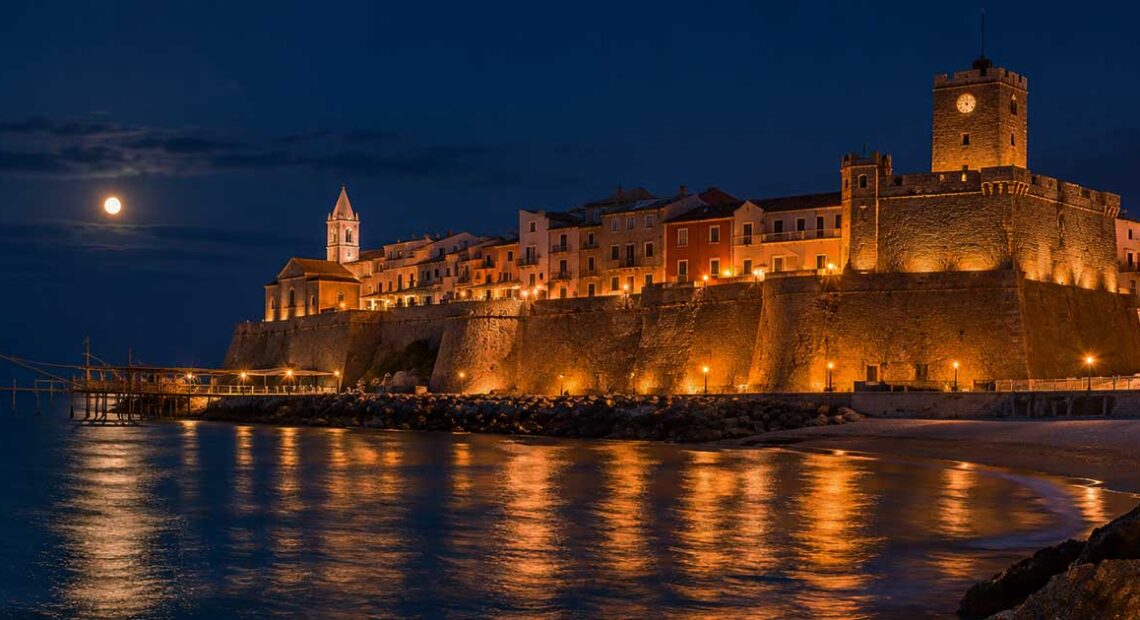 Suggestiva veduta notturna del Borgo Vecchio di Termoli illuminato con il Castello Svevo, il trabucco a mare e la luna piena che si riflette sull'acqua