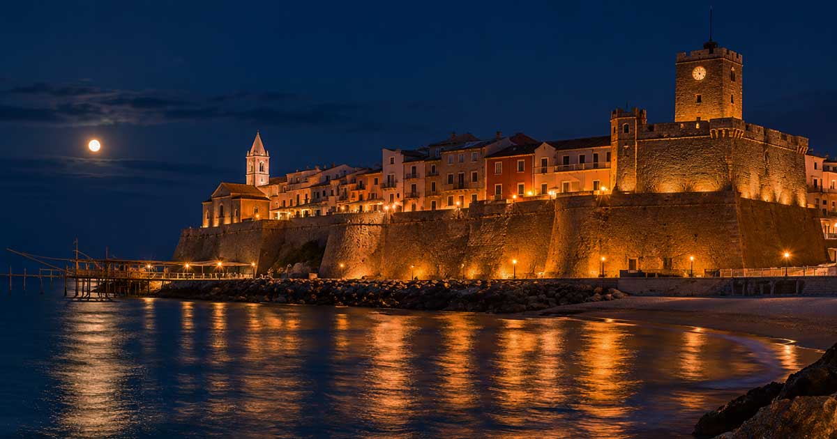 Suggestiva veduta notturna del Borgo Vecchio di Termoli illuminato con il Castello Svevo, il trabucco a mare e la luna piena che si riflette sull'acqua